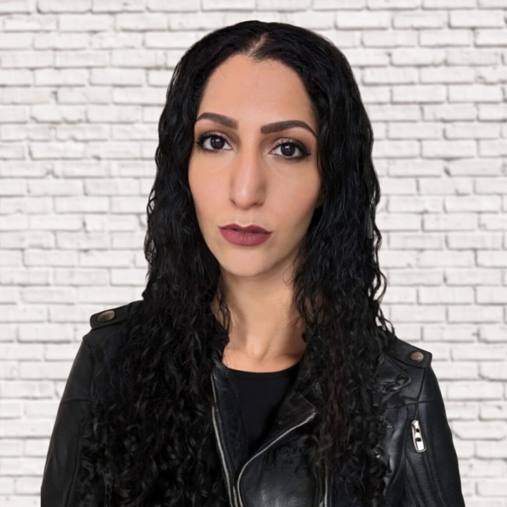 An image of Lilithfoxx wearing a black leather jacket and standing in front of white bricks.