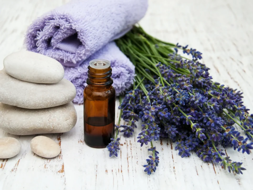 A bottle of massage oil, lavender flowers, massage stones, and a purple towel on a rustic white wooden table.