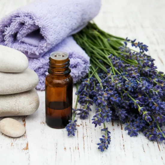 A bottle of massage oil, lavender flowers, massage stones, and a purple towel on a rustic white wooden table.